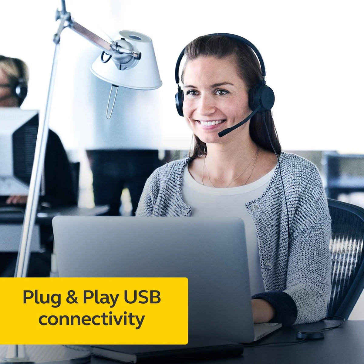 A woman with a headset, known for its all-day comfort and noise-canceling microphone, smiles while using a laptop in an office setting. A lamp is visible on her desk. Text overlay at the bottom-left corner reads "Plug & Play USB connectivity." The Jabra Evolve 20 UC Wired Headset Mono, Professional Telephone Headphones for Greater Productivity, Superior Sound for Calls and Music, USB Connection, All Day Comfort Design by JABRA ensures clarity as another person is blurred in the background.