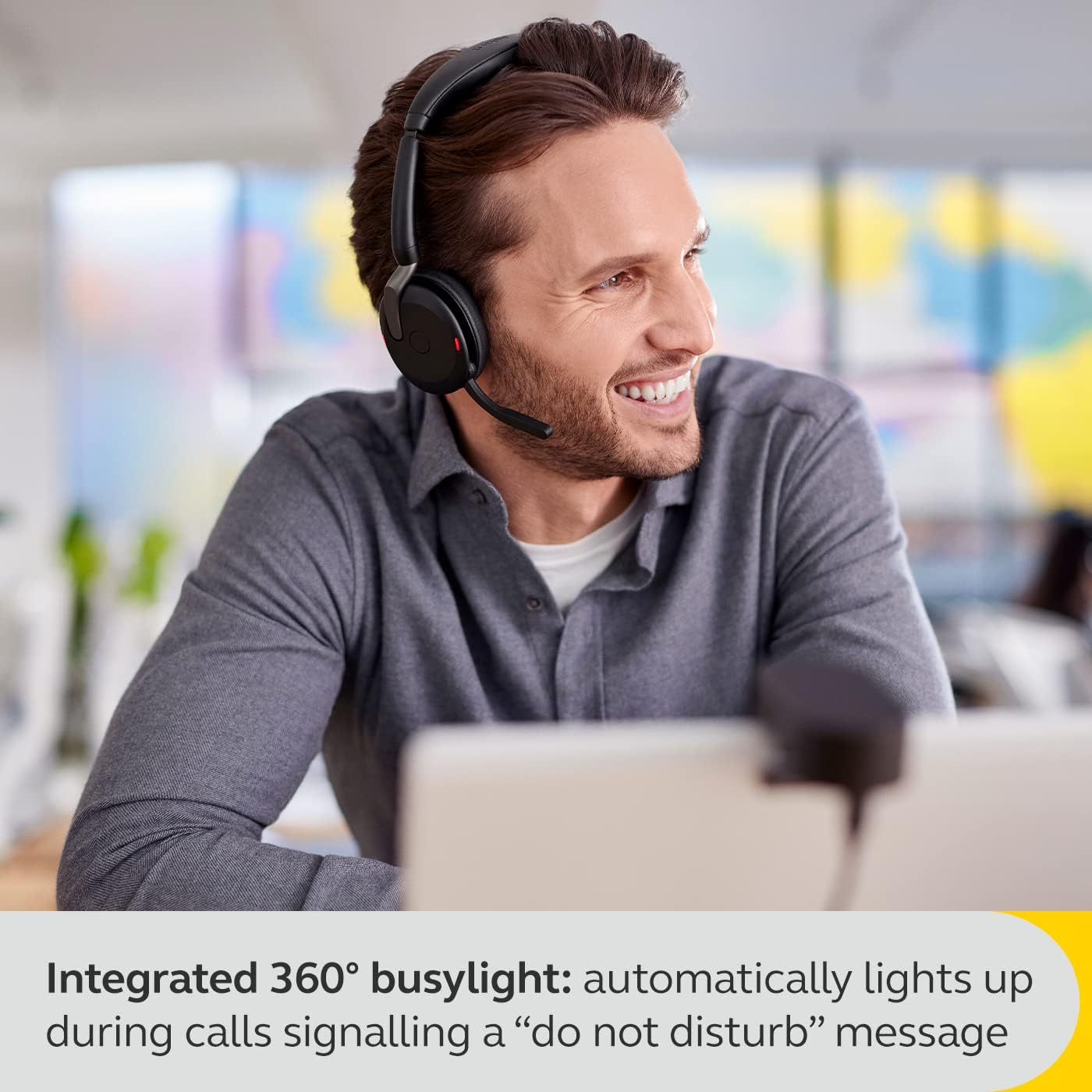 A smiling man is sitting at a desk, wearing a gray Jabra Evolve2 65 Flex Wireless Stereo Headset - Bluetooth, Noise-Cancelling ClearVoice Technology & Hybrid ANC - Works with All Leading UC Platforms Such As Zoom & Google Meet - Black with a microphone. The headset features an integrated 360° busylight that is lit up. The background shows a blurred office setting. Overlay text reads: "Integrated 360° busylight: automatically lights up during calls signaling a 'do not disturb' message.
