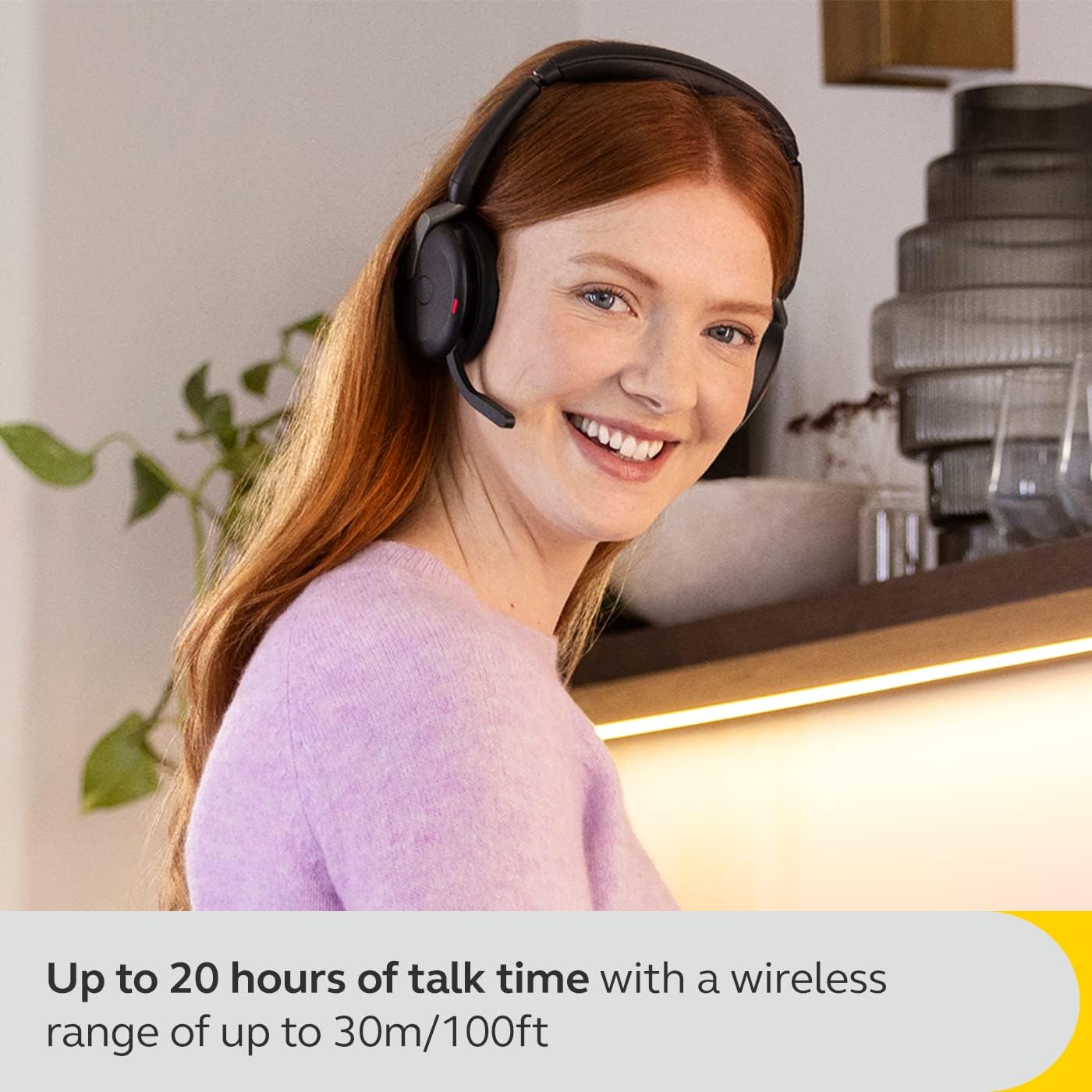 A woman with long red hair wearing a Jabra Evolve2 65 Flex Wireless Stereo Headset smiles while standing in a kitchen. Text on the image reads, "Up to 20 hours of talk time with Hybrid ANC and a wireless range of up to 30m/100ft.