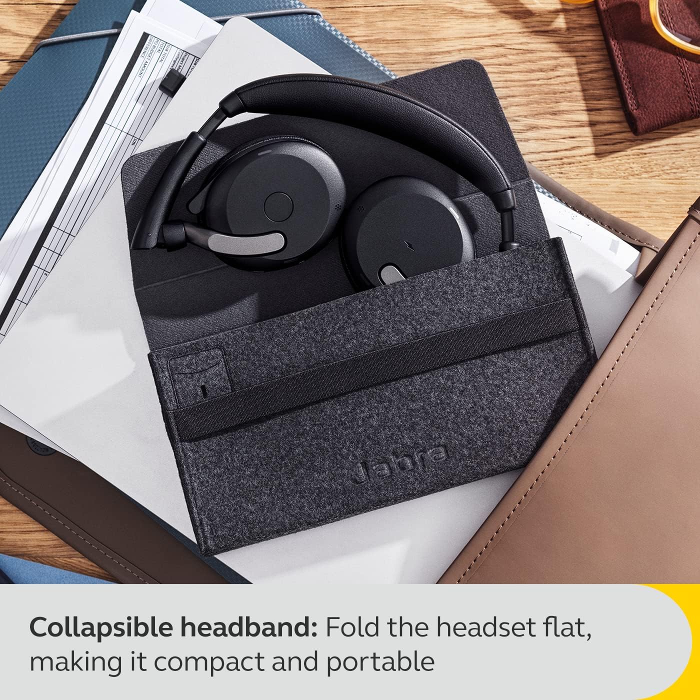 A pair of black JABRA Jabra Evolve2 65 Flex Wireless Stereo Headset - Bluetooth, Noise-Cancelling ClearVoice Technology & Hybrid ANC - Works with All Leading UC Platforms Such As Zoom & Google Meet is shown folded flat and placed in a gray felt carrying case. The case is resting among folders and papers on a wooden desk. The text reads: "Collapsible headband: Fold the headset flat, making it compact and portable.