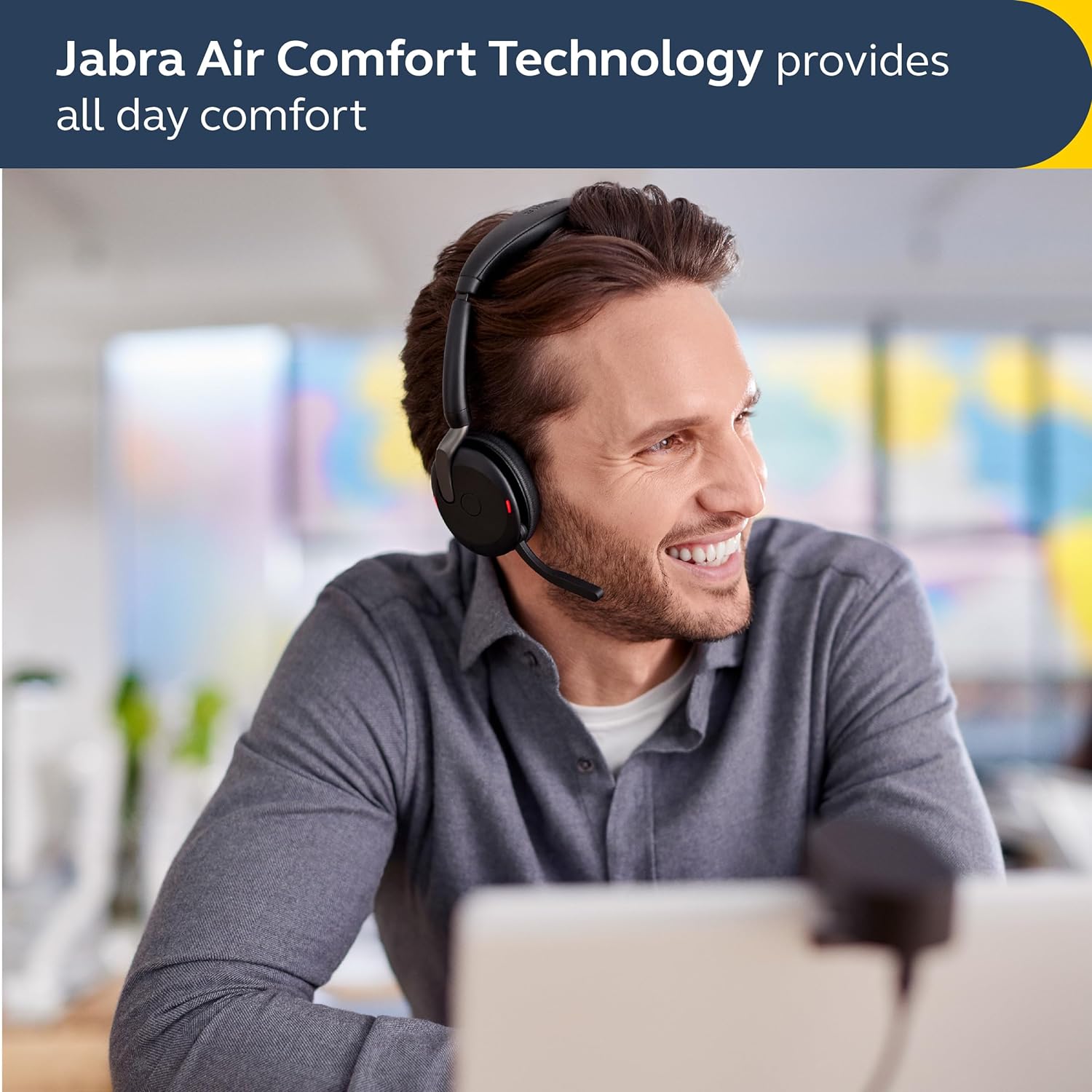 A man with short brown hair wearing a gray shirt and a headset smiles while looking to the side. He is seated in a modern, brightly lit office. The text "Jabra Evolve2 65 Flex Wireless Stereo Headset - Bluetooth, Noise-Cancelling ClearVoice Technology & Hybrid ANC - Works with All Leading UC Platforms Such As Zoom & Google Meet - Black provides all-day comfort" is displayed at the top.