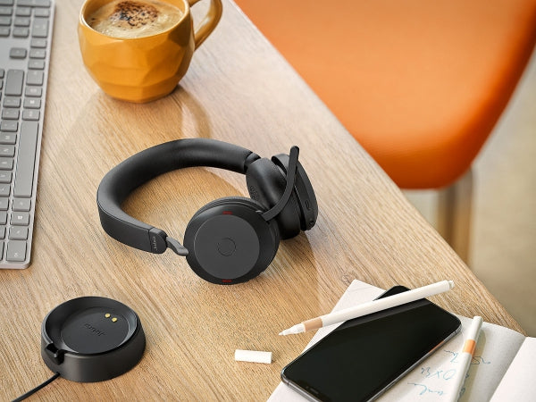 A workspace featuring a black headset with a microphone on a wooden desk. A charging dock, a smartphone, a notebook with pencils, a burrito, and a yellow coffee mug are also on the desk. Part of a keyboard and an orange chair are visible in the background.