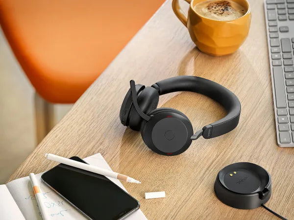 A work desk with black wireless headphones, a charging dock, a smartphone, a notebook with a pen, a cup of coffee, and a computer keyboard. The desk surface is wooden, and there is an orange chair in the background.