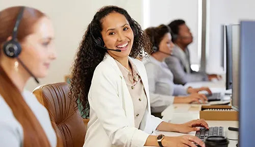 A woman with curly hair, wearing a white blazer and headset, smiles at the camera while typing on a keyboard. She is seated in an office environment with colleagues in the background, also wearing headsets and working on computers.