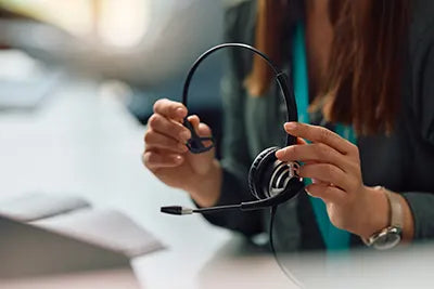 A person is sitting at a desk, holding a black headset with a microphone. They are partially visible from the neck down, wearing a teal shirt under a blazer. The background includes a computer keyboard and papers, suggesting an office environment.