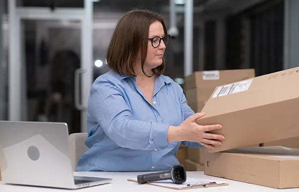 A woman wearing glasses and a blue shirt is sitting at a desk, packing a cardboard box. There are other boxes around her, a laptop to her left, and a barcode scanner in front of her. She appears focused on handling the package.