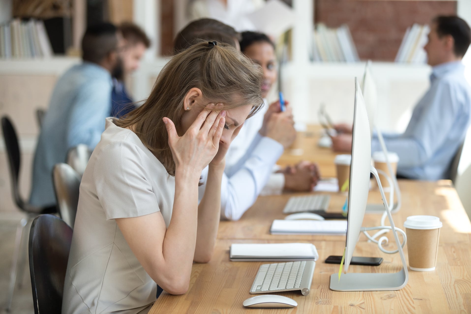 A woman with a headache sits at a desk in an office, holding her temples. She is in front of a computer, surrounded by other coworkers working and talking. The office has a modern design with wooden tables and shelves in the background.